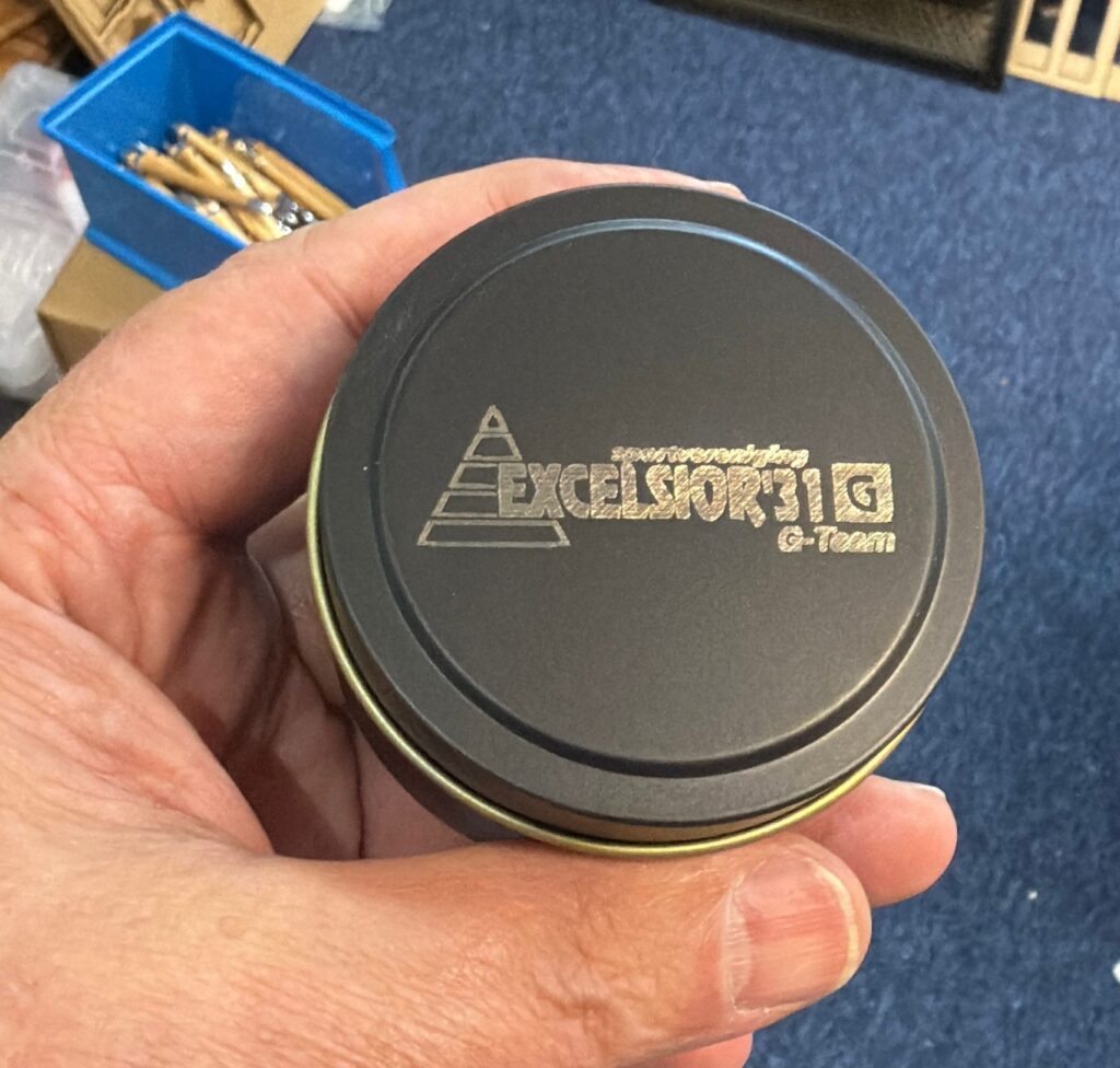 Hand houdt ronde metalen doos met Excelsior logo